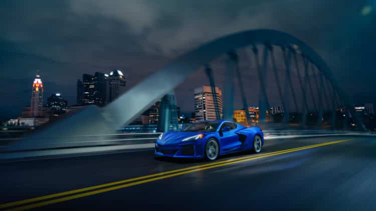 Front 3/4 view of 2024 Chevrolet Corvette E-Ray 3LZ coupe in Riptide Blue, driving over a bridge in front of a city. Pre-production model shown. Actual production model may vary. Model year 2024 Corvette E-Ray available 2023.