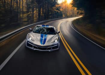 Front view of 2024 Chevrolet Corvette E-Ray 3LZ convertible in Silver Flare with Electric Blue stripe package driving on a road between trees. Pre-production model shown. Actual production model may vary. Model year 2024 Corvette E-Ray available 2023.