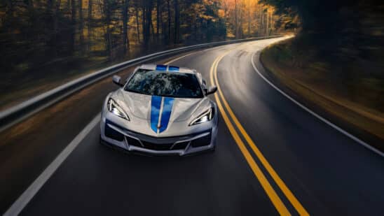 Front view of 2024 Chevrolet Corvette E-Ray 3LZ convertible in Silver Flare with Electric Blue stripe package driving on a road between trees. Pre-production model shown. Actual production model may vary. Model year 2024 Corvette E-Ray available 2023.