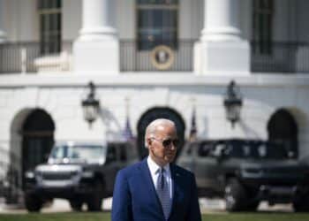 President Biden standing with the White House far behind him, as well as vehicles parked behind him