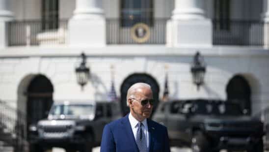 President Biden standing with the White House far behind him, as well as vehicles parked behind him