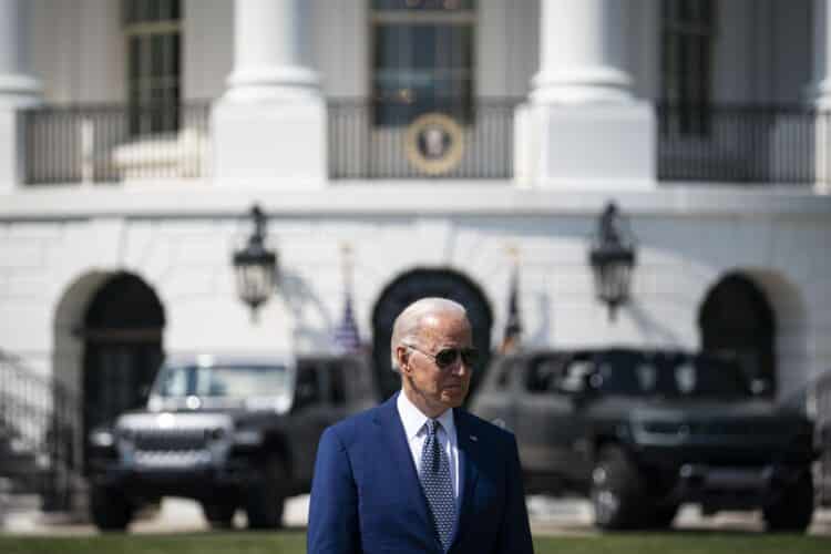 President Biden standing with the White House far behind him, as well as vehicles parked behind him